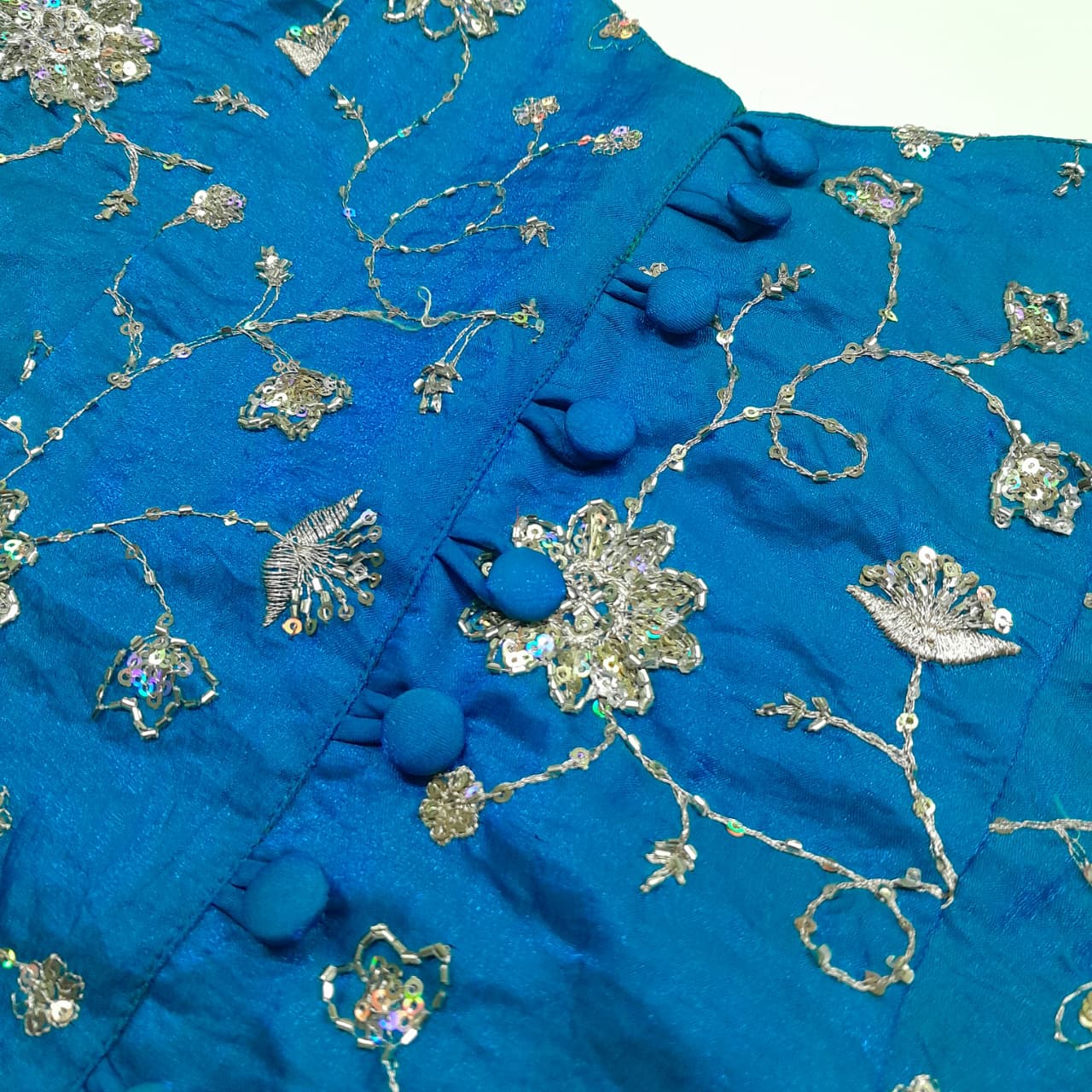 A close-up of a sky blue corset featuring intricate gold metallic embroidery, sequins, and decorative button details.