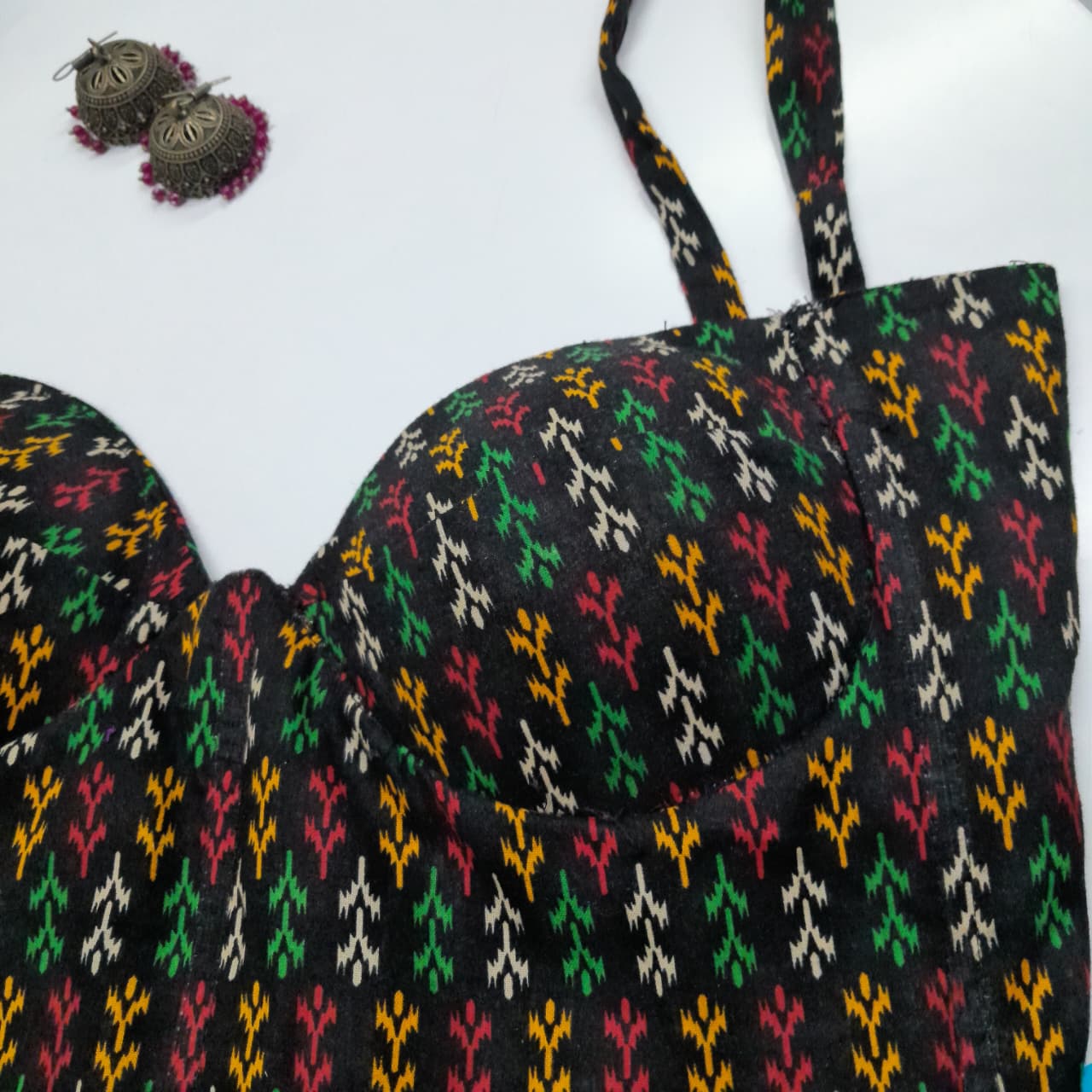 A close-up of a black bralette corset with a colorful ikat pattern in green, red, yellow, and white, featuring molded cups and thin straps, shown on a white background with a pair of earrings in the top-left corner.
