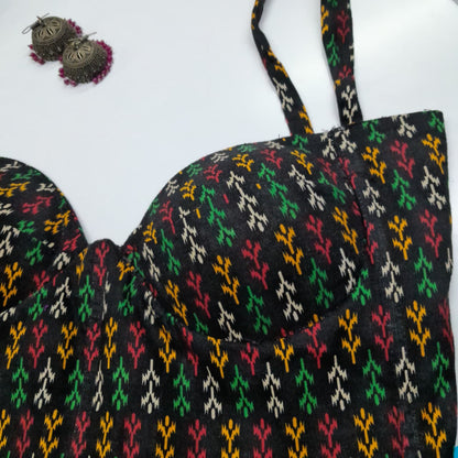 A close-up of a black bralette corset with a colorful ikat pattern in green, red, yellow, and white, featuring molded cups and thin straps, shown on a white background with a pair of earrings in the top-left corner.