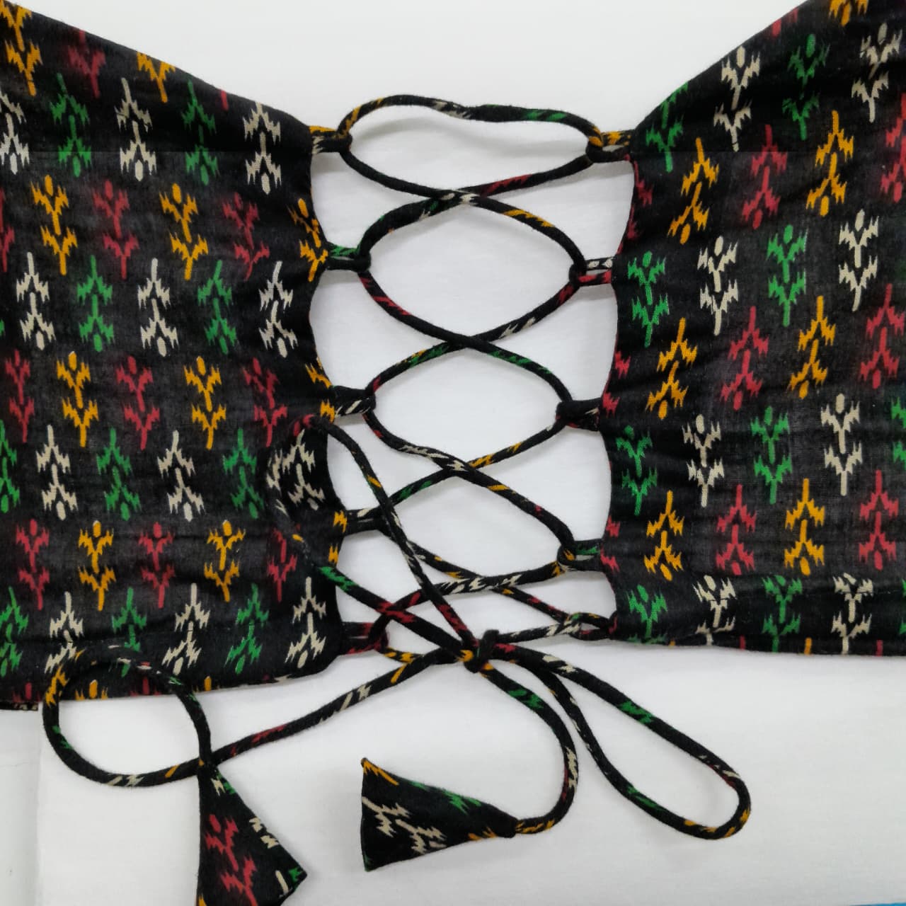 A close-up of a black lace-up bustier corset with a colorful ikat pattern in green, red, yellow, and white, tied with matching black cord on a white background.