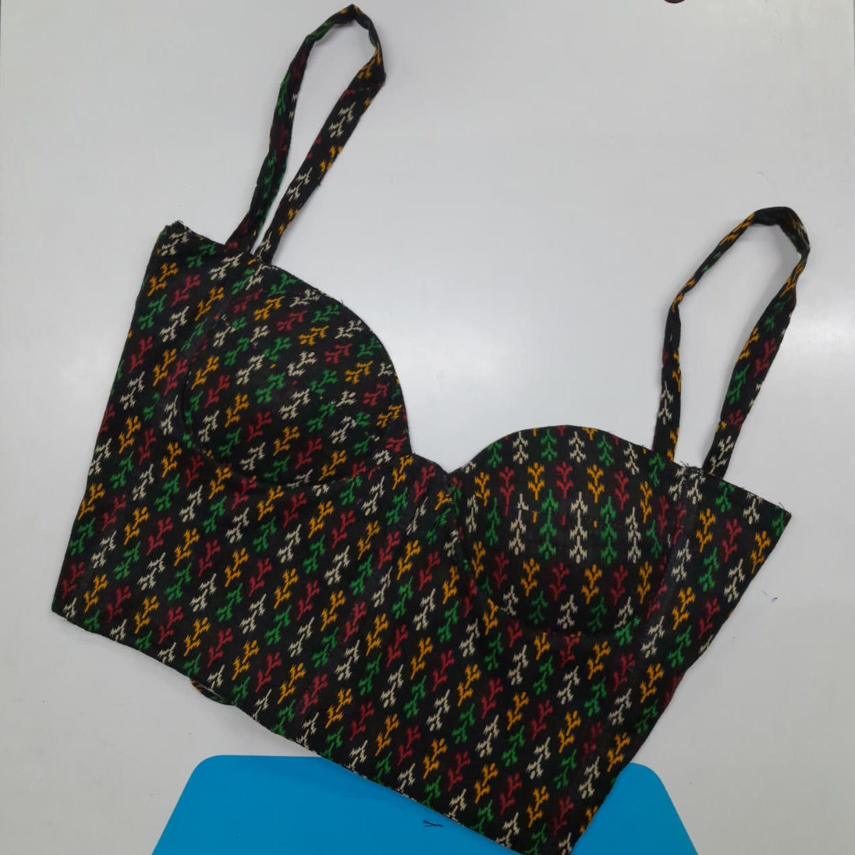 A bustier-style bra top with a multicolor geometric pattern on a black base, featuring thin shoulder straps, laid flat on a white surface.