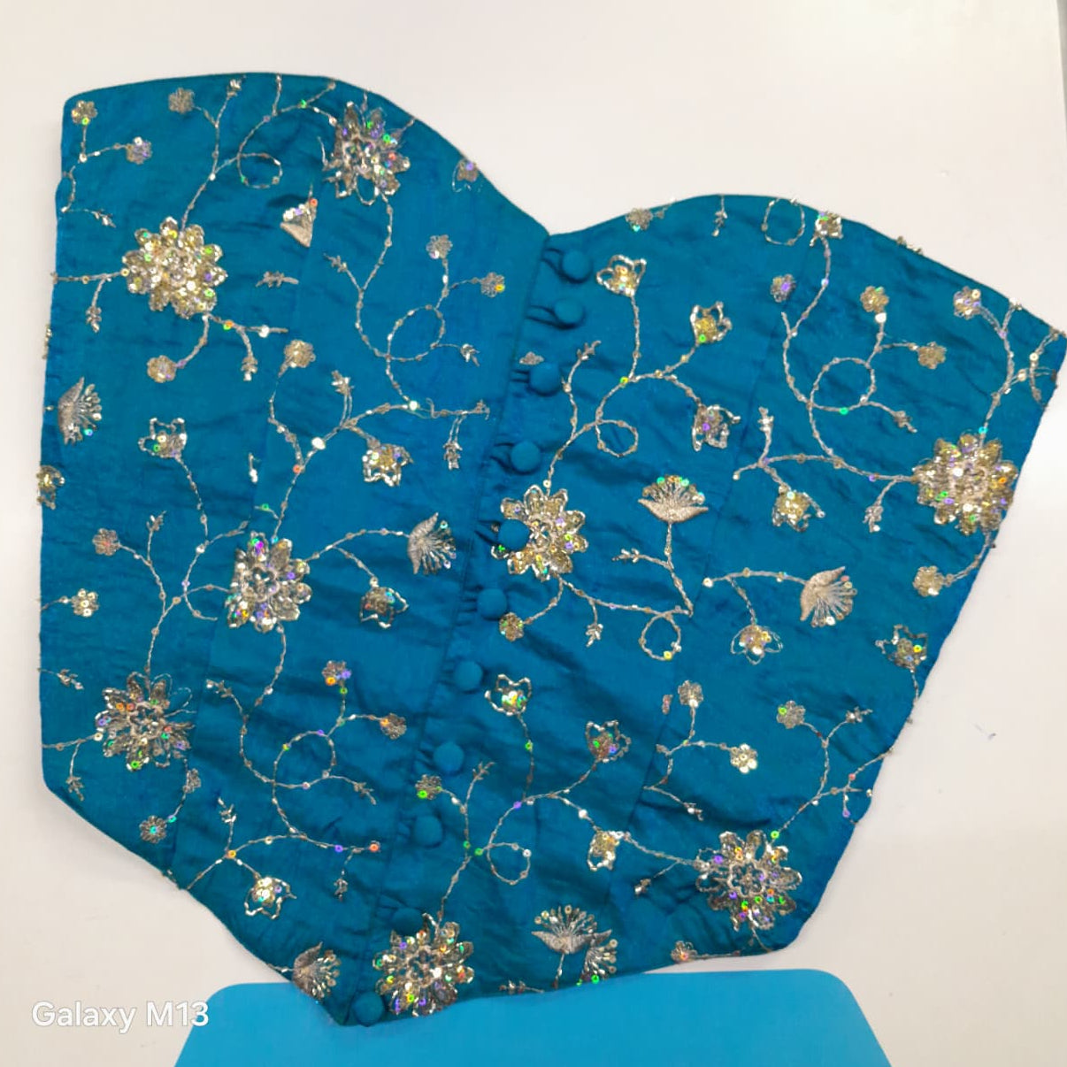 A sky blue strapless corset with intricate gold zari embroidery, floral patterns, and rhinestone embellishments, laid flat on a white surface.